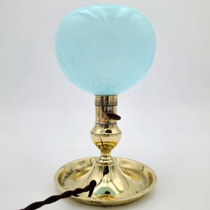 Puede incluir: Una lámpara de mesa vintage con una pantalla de globo de cristal azul claro. La lámpara tiene una base de latón y un cable eléctrico marrón. La base incluye un plato redondo y un tallo decorativo. La lámpara está diseñada para una mesa.