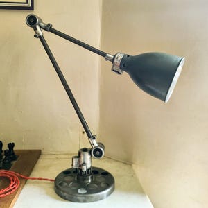 Puede incluir: Lámpara de escritorio ajustable con una pantalla gris oscuro y un brazo articulado plateado. La lámpara tiene una base circular con múltiples agujeros y un cable de alimentación rojo. Se encuentra sobre una superficie blanca contra una pared clara.