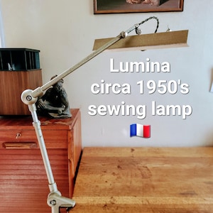 Könnte beinhalten: Eine Vintage Lumina Nählampe aus den 1950er Jahren. Die Lampe hat einen silbernen, verstellbaren Arm und eine rechteckige Leuchte. Die Lampe ist an einem Holztisch befestigt. Der Text "Lumina circa 1950's sewing lamp" ist sichtbar.