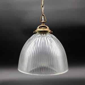 Peut inclure: Suspension luminaire en verre transparent avec un abat-jour en forme de dôme nervuré. Le luminaire a un capuchon et une chaîne en métal de couleur laiton. La lumière est suspendue à une chaîne sur fond sombre.