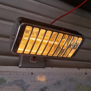 May include: A vintage Thermor electric heater with a chrome finish and a rectangular design. The heater has a series of vertical bars across the front and a red power cord. The Thermor logo is visible on the base. The heater is mounted on a wall.