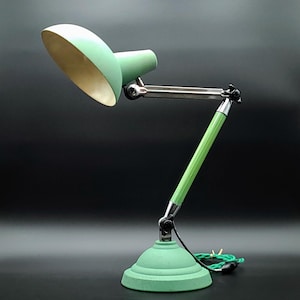 May include: A mint green desk lamp with a rounded base and adjustable arm. The lamp shade is mint green on the outside and gold on the inside. The lamp has a silver-coloured arm and a green power cord.