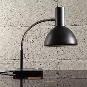 May include: A black desk lamp with a flexible, silver gooseneck arm and a dome-shaped shade. The lamp sits on a rectangular base with a switch and a power cord. The background is a textured white brick wall.