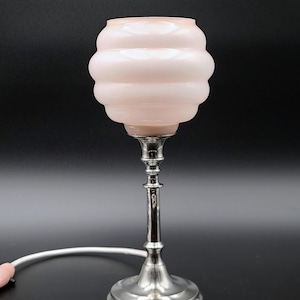 Puede incluir: Una pequeña lámpara de mesa vintage con una pantalla de cristal rosa acanalado y una base de metal plateado. La lámpara tiene un tallo largo y delgado y una base redonda. El cable de alimentación es blanco.