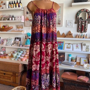 Könnte beinhalten: Ein bodenlanges Kleid mit dünnen Trägern, ausgestellt auf einer Schaufensterpuppe. Das Kleid hat ein mehrschichtiges Design mit Farben wie Rot, Lila und Orange sowie ein Blumenmuster. Das Kleid ist aus leichtem Stoff gefertigt.