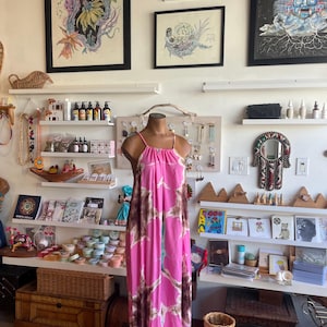 Könnte beinhalten: Ein pinkfarbenes und braunes Maxikleid mit Muster auf einer Schaufensterpuppe. Das Kleid hat einen Neckholder-Ausschnitt und eine fließende Silhouette. Der Hintergrund zeigt gerahmte Kunstwerke, Regale mit verschiedenen Produkten und Dekorationsgegenständen.