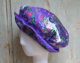 May include: A purple felt beret with a floral pattern in red, green, and white. The hat is displayed on a white mannequin head. The beret has a rounded crown and a small brim, showcasing a unique, handcrafted design.