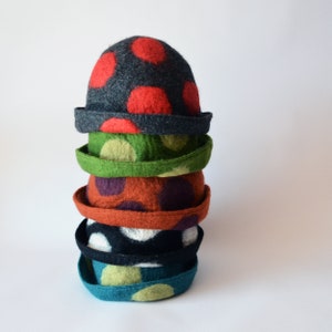 Puede incluir: Una pila de seis sombreros de fieltro de lana con lunares de diferentes colores. Los sombreros están apilados del más grande al más pequeño, con el sombrero superior siendo gris con lunares rojos. Los sombreros están todos hechos de fieltro y tienen una textura suave y peluda.