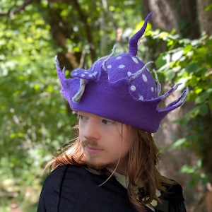 Pode incluir: Um chapéu de feltro roxo vibrante com um topo pontiagudo e múltiplas protuberâncias curvas semelhantes a tentáculos que se estendem da aba. A superfície do chapéu é decorada com pontos circulares brancos e detalhes cinzentos texturizados.