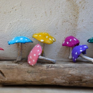 May include: A collection of eight felt mushrooms in various colors, including pink, blue, yellow, green, and purple, with white polka dots. The mushrooms are arranged on a piece of driftwood.