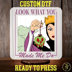 May include: A square, light-colored wooden sign featuring a cartoon image of the Evil Queen from Snow White holding a green, skull-shaped apple. Text includes "CUSTOM DTF", "LOOK WHAT YOU", "Made Me Do", and "READY TO PRESS".