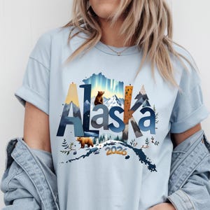 May include: Light blue t-shirt with the word "Alaska" in a stylized design featuring mountains, trees, and bears. The year "2026" is printed below the word. The shirt is made of a soft material.