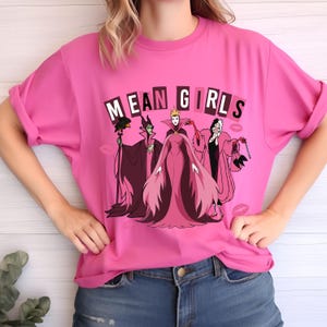 May include: A bright pink t-shirt with the words "MEAN GIRLS" above a cartoon graphic of three villains. The villains are in pink, black, and purple. The shirt is worn with blue jeans.
