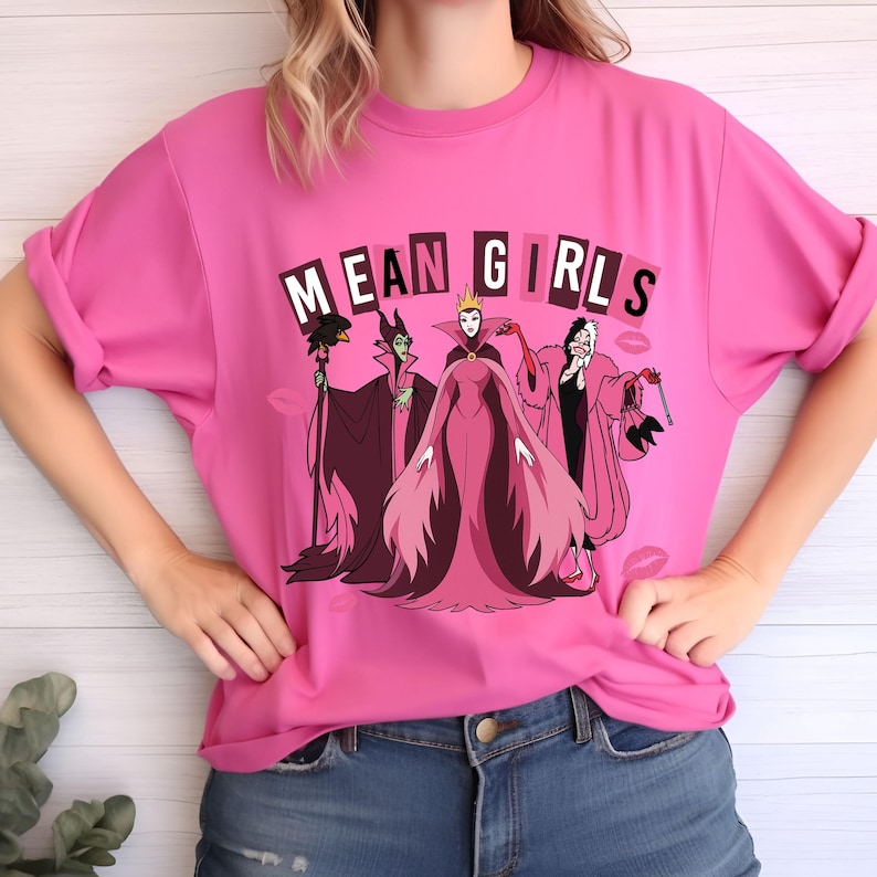 May include: A bright pink t-shirt with the words "MEAN GIRLS" above a cartoon graphic of three villains. The villains are in pink, black, and purple. The shirt is worn with blue jeans.