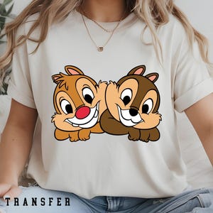 May include: A cream-coloured t-shirt with a graphic of two cartoon chipmunks, one with a red nose. The chipmunks are smiling and lying down. The text "DTF TRANSFER" is printed at the bottom of the shirt.