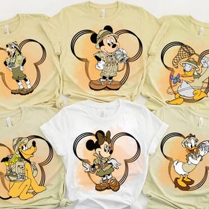 May include: Set of six t-shirts featuring different Disney characters in safari outfits. Each shirt has a cartoon character wearing a safari hat and holding a safari-themed item, such as binoculars or a net. The shirts are all beige with a brown Mickey Mouse silhouette.