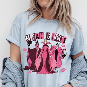 May include: Pale blue t-shirt with a graphic of four cartoon villains and the text "MEAN GIRLS" in pink and white. The design includes pink lipstick kiss marks.
