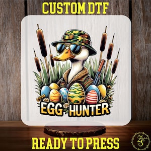 May include: A cartoon duck wearing camouflage and sunglasses, holding a gun and surrounded by colorful Easter eggs. The text "Egg Hunter" is below the duck.