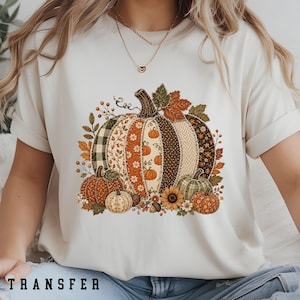 May include: A cream-coloured short-sleeve t-shirt featuring a large central graphic of a patchwork-style pumpkin surrounded by autumn leaves, smaller pumpkins, and a sunflower. The design uses various patterns including plaid, floral, and polka dots.