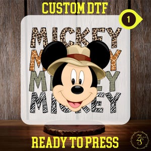 May include: A white heat transfer design featuring Mickey Mouse wearing a safari hat. The design includes the word "Mickey" repeated four times, each letter covered in a different animal print: leopard, giraffe, crocodile, and zebra.