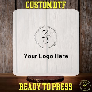 May include: A white, rectangular, blank transfer sheet with a black circle containing a stylized letter "Z" with a line through it and the text "D.T.F." above it. The text "Your Logo Here" is below the circle. The sheet is on a wooden surface with the text "CUSTOM DTF" above it and "READY TO PRESS" below it.