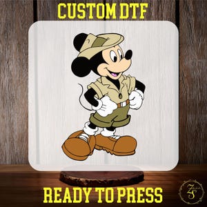 May include: A cartoon image of Mickey Mouse wearing a safari hat and outfit. The image is ready to be pressed onto a t-shirt or other fabric.