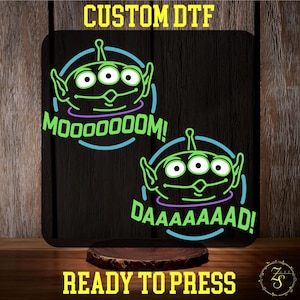 May include: Two green alien head designs with three eyes and a mouth, on a black background. The top design has the text "MOOOOOM!" and the bottom design has the text "DAAAAAAAAAD!"  The text is in a neon blue colour.  The background is black.  The text "CUSTOM DTF READY TO PRESS" is at the top and bottom of the image.