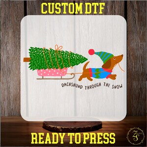May include: A white square featuring a festive graphic. A dachshund dog in a blue and green sweater pulls a pink sled with a green Christmas tree. Text reads "DACHSHUND THROUGH THE SNOW". The top text says "CUSTOM DTF" and the bottom text says "READY TO PRESS".