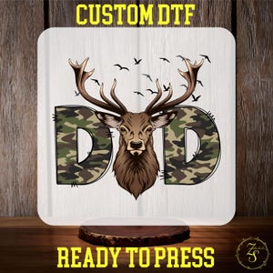 May include: A white background with a camouflage pattern "D" on either side of a brown deer head with antlers. The text "CUSTOM DTF" is at the top of the image and "READY TO PRESS" is at the bottom.