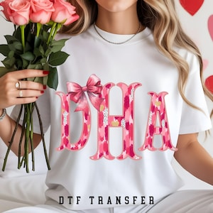 May include: White t-shirt with the letters DHA in pink and red watercolor-style design, accented with a pink bow. The text "DTF TRANSFER" is printed at the bottom. A bouquet of pink roses is held in the image.