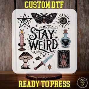 May include: A white background with a black and white design featuring a moth, a candle, a mushroom, a dagger, a tarot card, a potion bottle, a star with an eye, and a dice. The text "Stay Weird" is in black lettering.