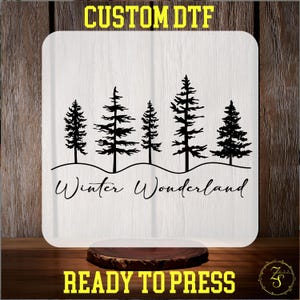 May include: A white square with rounded corners features a black silhouette of pine trees and the words "Winter Wonderland." The text "CUSTOM DTF" and "READY TO PRESS" are in yellow. The design is on a wooden surface.