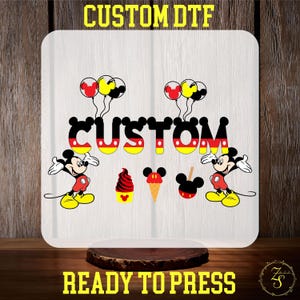 May include: A white iron-on transfer design featuring Mickey Mouse characters, balloons, ice cream, and the word "CUSTOM" in a Mickey Mouse font. The design is ready to press.