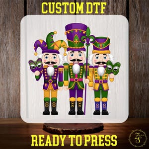May include: Three nutcracker figurines in Mardi Gras colors. The nutcrackers are wearing purple, green, and gold costumes. The middle nutcracker is holding a green staff with a fleur-de-lis on top. The nutcracker on the left is wearing a jester hat and holding a purple and green mask. The nutcracker on the right is holding a gold and green mask. The text "CUSTOM DTF" is at the top of the image and "READY TO PRESS" is at the bottom.