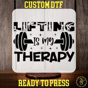 May include: A white square sign with the text "LIFTING IS MY THERAPY" in black, featuring dumbbell illustrations and star accents. The sign also includes the text "CUSTOM DTF" and "READY TO PRESS" in yellow.