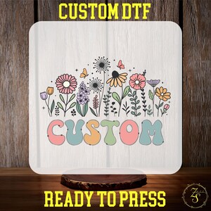 May include: A square DTF transfer sheet featuring a colorful floral illustration with pink, orange, purple, and green flowers, dandelions, and butterflies. The word CUSTOM is printed in large, rounded, multi-colored bubble letters below the flowers.