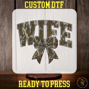 May include: A white iron-on transfer featuring the word "WIFE" in a camouflage pattern with a bow in the centre. The transfer is ready to press.