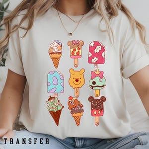 May include: Cream-colored t-shirt featuring colorful cartoon ice cream cones and popsicles. Designs include ice cream cones and character-themed popsicles. The text "DTF TRANSFER" is printed at the bottom of the shirt.