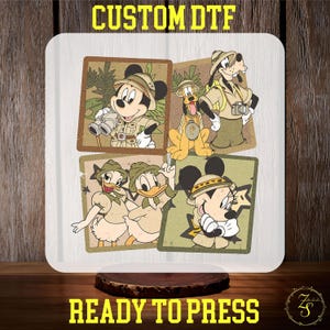 May include: A white square design transfer sheet with four images of Disney characters dressed in safari outfits. The characters include Mickey Mouse, Minnie Mouse, Donald Duck, and Goofy. The images are surrounded by a green and brown patterned border.