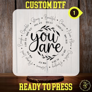 Peut inclure: Fond blanc avec un cercle noir contenant les mots "you are" dans une police décorative. Le cercle est entouré d'une couronne de feuilles. Autour du cercle se trouvent des mots comme "Strong", "Beautiful", "Chosen", "Never Alone", "Created", "Amazing", "Victorious", "Always Loved", "Enough", "Capable". Chaque mot est accompagné d'un cœur et d'un verset biblique.