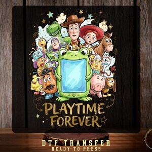 May include: A DTF transfer sheet featuring a colorful illustration of various animated toy characters surrounding a central green frog-shaped frame. Below the characters, the text PLAYTIME FOREVER is printed in a gold, textured font style.