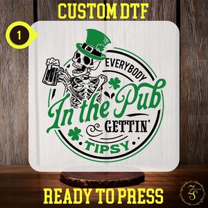 May include: A white, round, iron-on transfer design featuring a skeleton wearing a green hat and holding a beer mug. The design says "Everybody In the Pub & Gettin' Tipsy."