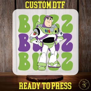 May include: A white heat transfer design featuring a cartoon image of Buzz Lightyear from the Toy Story franchise. The design includes the word "Buzz" repeated three times in a retro font. The design is ready to press.