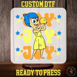 May include: A white background with a yellow, orange, and blue design featuring a cartoon character holding a yellow ball. The text "Joy" is written in a large, stylized font. The design is surrounded by blue and orange stars. The text "Custom DTF Ready to Press" is written below the design.