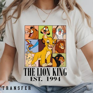 May include: Cream-colored t-shirt featuring a colorful graphic of characters from The Lion King, including Simba, Timon, and Pumbaa. The text "THE LION KING EST. 1994" is printed below the graphic. The shirt is a DTF transfer.