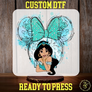 May include: A white square with a cartoon illustration of Princess Jasmine, a teal bow, and Mickey Mouse ears. The text "CUSTOM DTF" and "READY TO PRESS" are in yellow. The image is on a wooden surface.