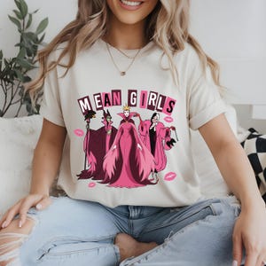 May include: A cream-coloured t-shirt with the words "MEAN GIRLS" above a graphic of three cartoon villains in pink and purple robes. Pink lipstick kiss marks are scattered around the graphic.
