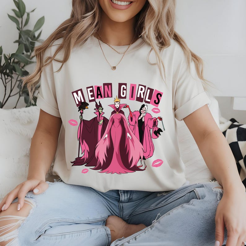 May include: A cream-coloured t-shirt with the words "MEAN GIRLS" above a graphic of three cartoon villains in pink and purple robes. Pink lipstick kiss marks are scattered around the graphic.