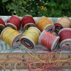 May include: A collection of 10 wooden spools of thread in various shades of yellow, orange, and red. The spools are labeled "COATS & CLARK'S" and "CLARK'S" and are arranged on a white and green patterned surface.