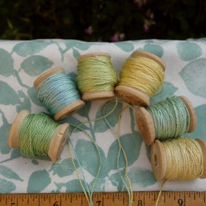 May include: Six wooden spools of embroidery thread in shades of light blue, sage green, and pale yellow, arranged in a semi-circle. A wooden ruler at the bottom shows measurements up to 10 inches, indicating the scale of the thread spools.